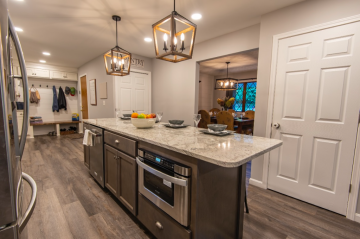 A kitchen with an island that has a stove, dishwasher, and cabinets on one site and barstools on the other side. In the background is the pantry, entryway, and dinning room showing the construction skills of ATM Contracting LLC.