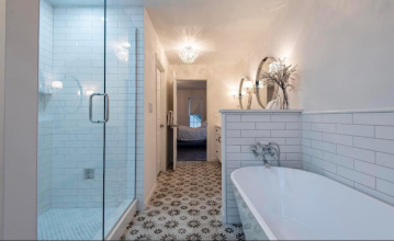 A bathroom with a shower that has glass doors, small tile on the floor and the wall with larger titles, decorative title on the floor of the bathroom, double sinks, and a vintage styled bathtub with tile on the wall around the tub showing the tile work fo